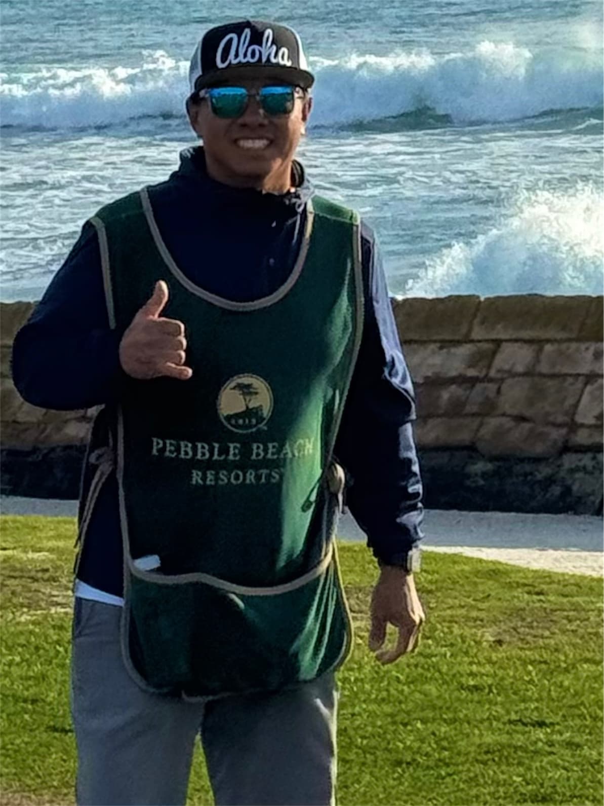 Johnny Eugenio in his caddie bib giving a shaka by the seawall, with surf breaking behind him