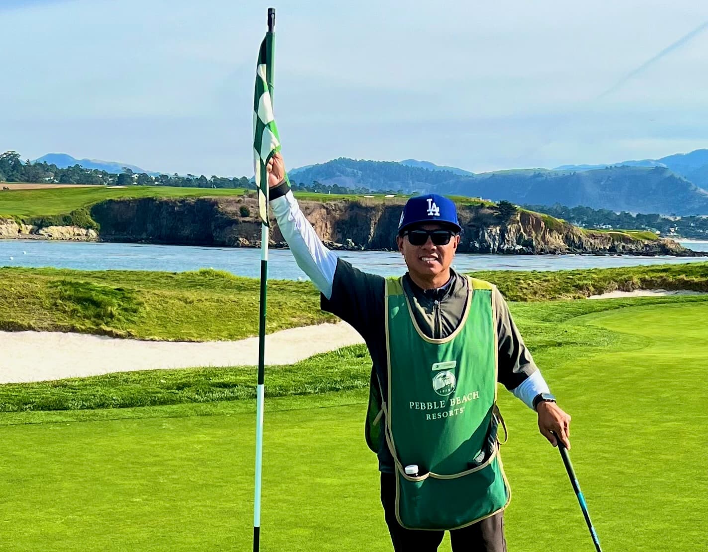 Johnny holding the flagstick on a green above the bay, with cliffs and mountains behind