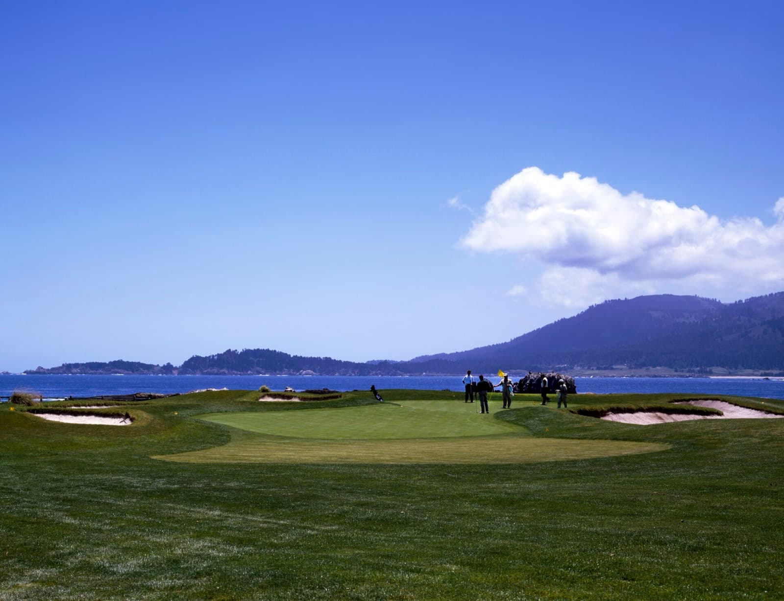 A green above Carmel Bay at Pebble Beach Golf Links, with golfers and the coastline behind