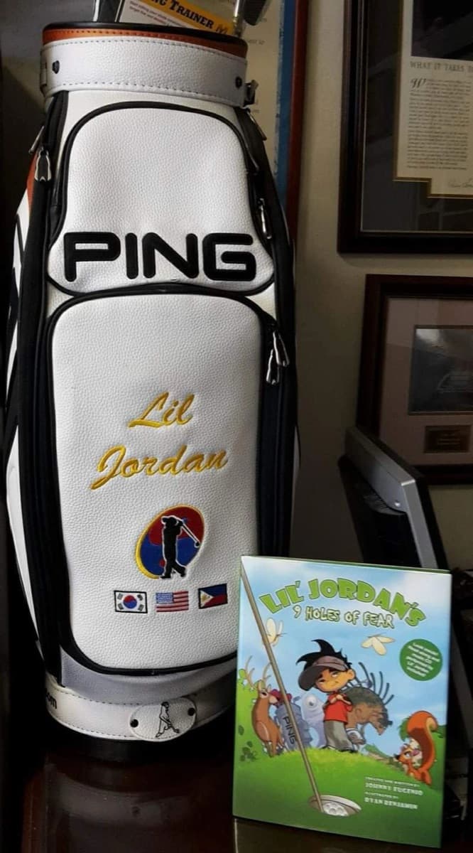 A PING golf bag with Lil’ Jordan on it, next to the book